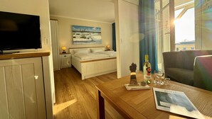 Une chambre d’hôtel avec un lit, une télévision, un tableau au mur, une table avec un journal et une fenêtre avec des rideaux.