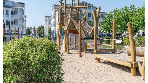 Eine hölzerne Spielgeräte-Konstruktion mit einer Seilbrücke und einer Rutsche, umgeben von Sand und Grünfläche.