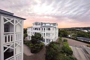 A white building with a balcony, surrounded by greenery and a paved area. - Seepark Sellin Ferienwohnung nahe Strand (Sellin)