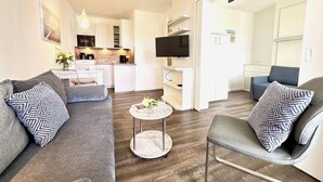 A modern living room with a sofa, armchair, and coffee table, a kitchen in the background, and a TV mounted on the wall. - Urlaub im Seepark Sellin (Sellin)