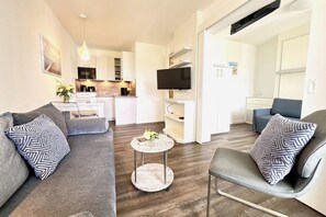 Ein modernes Wohnzimmer mit Sofa, Sessel und Couchtisch, im Hintergrund eine Küche und ein an der Wand befestigter Fernseher.