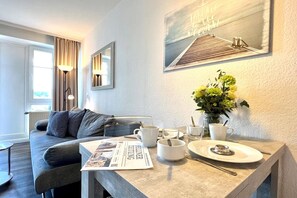 Ein modernes Wohnzimmer mit einer Couch, einem Esstisch mit einer Zeitung, einer Vase mit Blumen und einem Bild an der Wand.