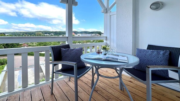 A covered outdoor seating area with a table and chairs, overlooking a landscape with greenery and buildings. - Urlaub im Seepark Sellin (Sellin)