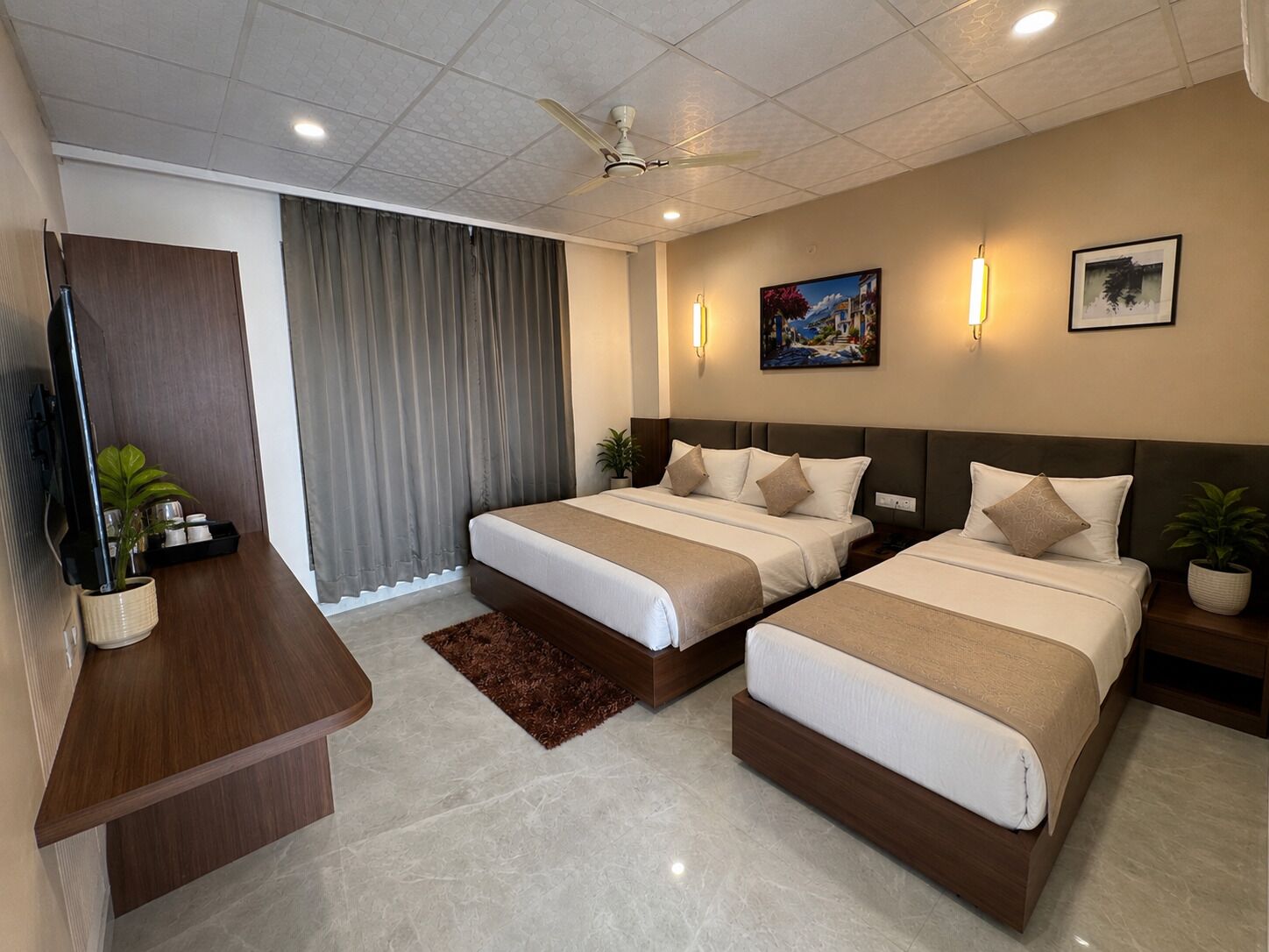 A hotel room with two beds, a desk, a ceiling fan, and a large window with curtains.