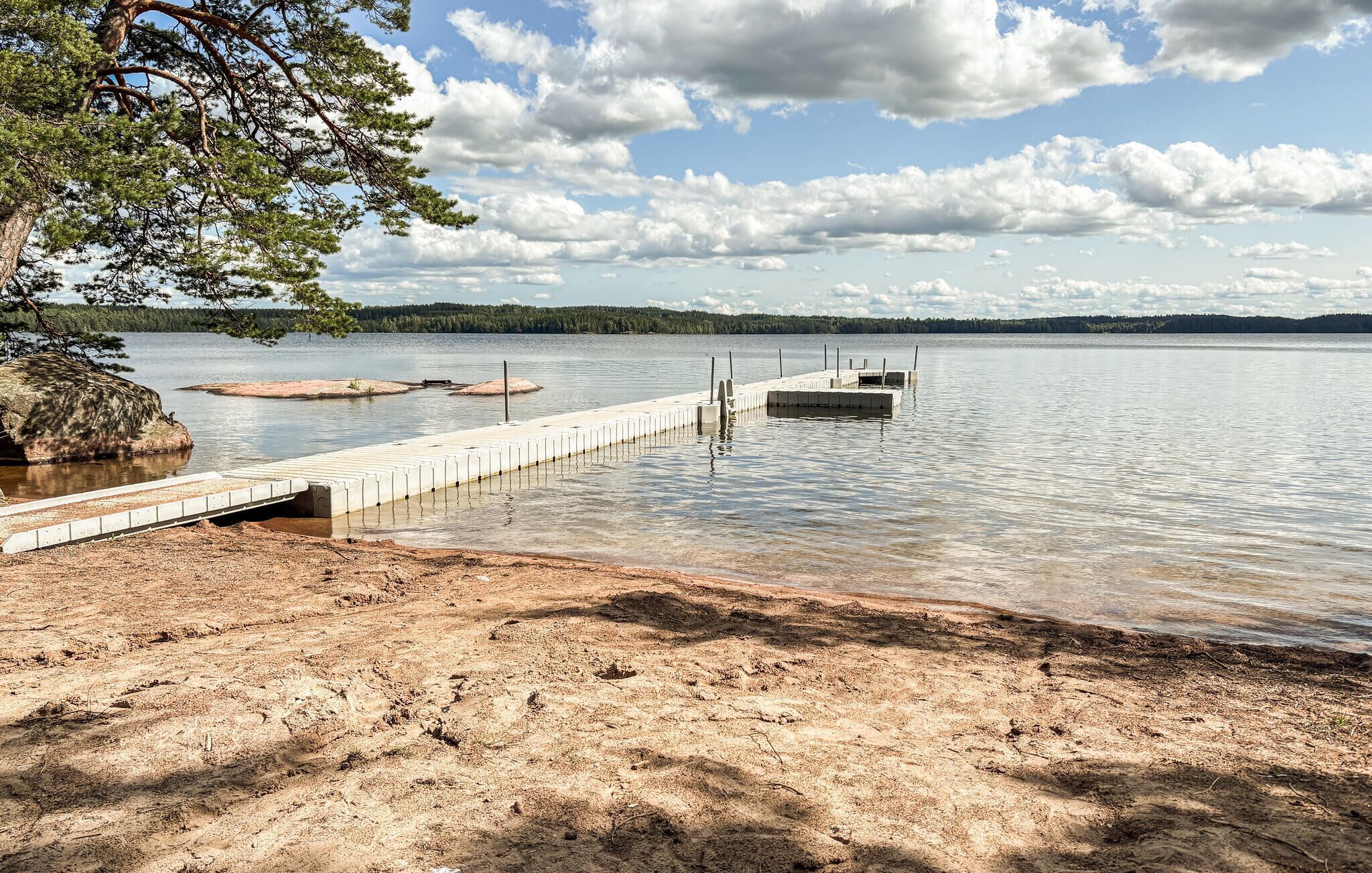 Un lac avec une jetée en béton qui s’avance dans l’eau, bordé d’une plage de sable et entouré d’arbres.
