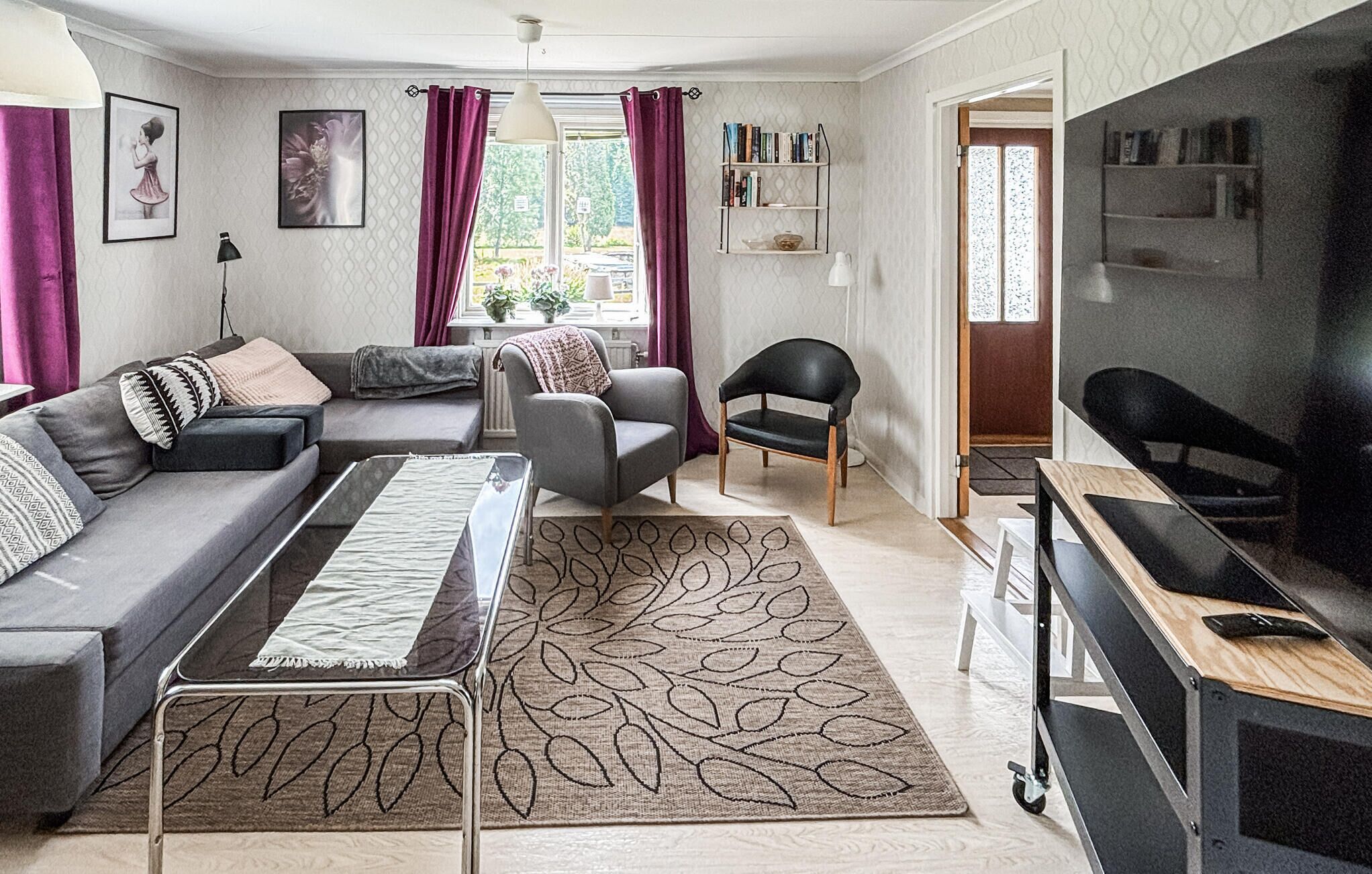 Una sala moderna con un sofá, un sillón y un escritorio con una silla. Se ve una estantería y una ventana con cortinas.