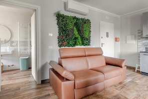 A modern living room with a brown leather sofa, a vertical garden wall, and a kitchen area in the background. - Residence Maratta 54 Orchidea (Terni)