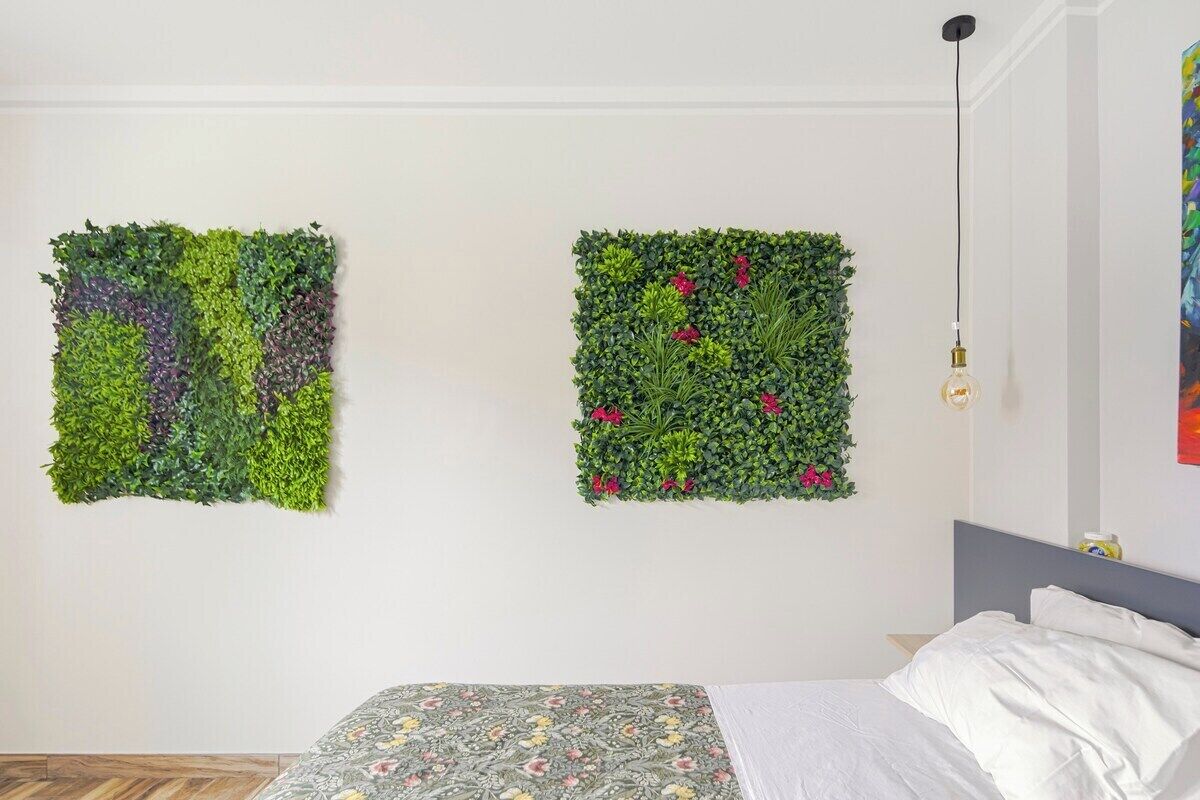 Ein Zimmer mit einem Bett, einer Wandleuchte und zwei quadratischen Wanddekorationen mit Grünpflanzen und Blumen.