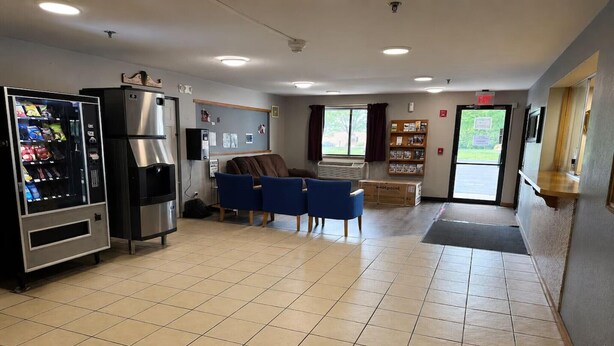 A clean, well-lit indoor space with a vending machine, a refrigerator, blue chairs, a small table, and a door leading outside. - 2 Full Beds | Non-Smoking (Coldwater, MI)