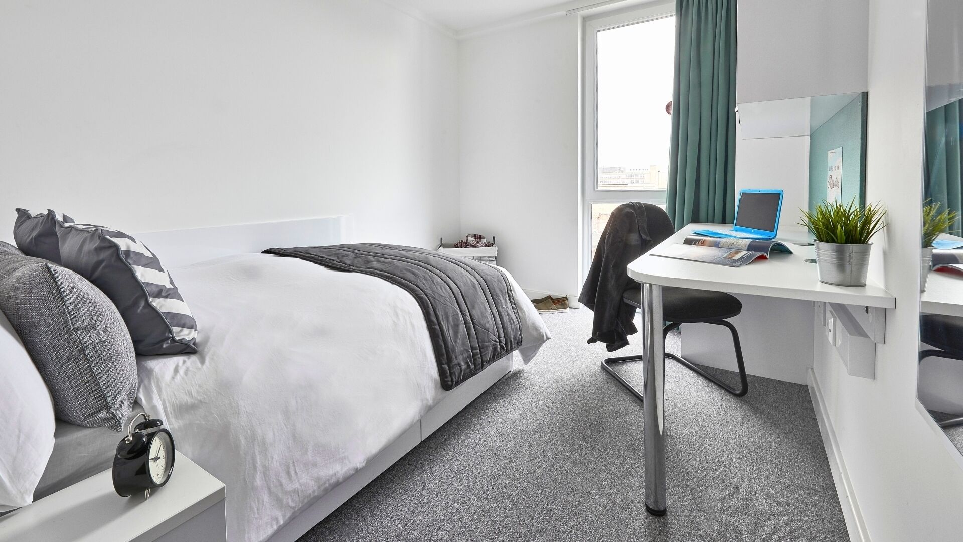 Hotelkamer met een bed, bureau, stoel, kleine tafel, een potplant en een raam met gordijnen.