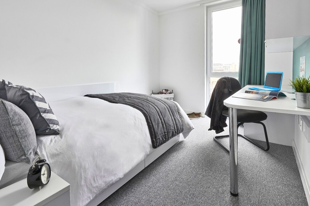 Ein Hotelzimmer mit einem Bett, einem Schreibtisch, einem Stuhl, einem kleinen Tisch, einer Zimmerpflanze und einem Fenster mit Vorhängen.
