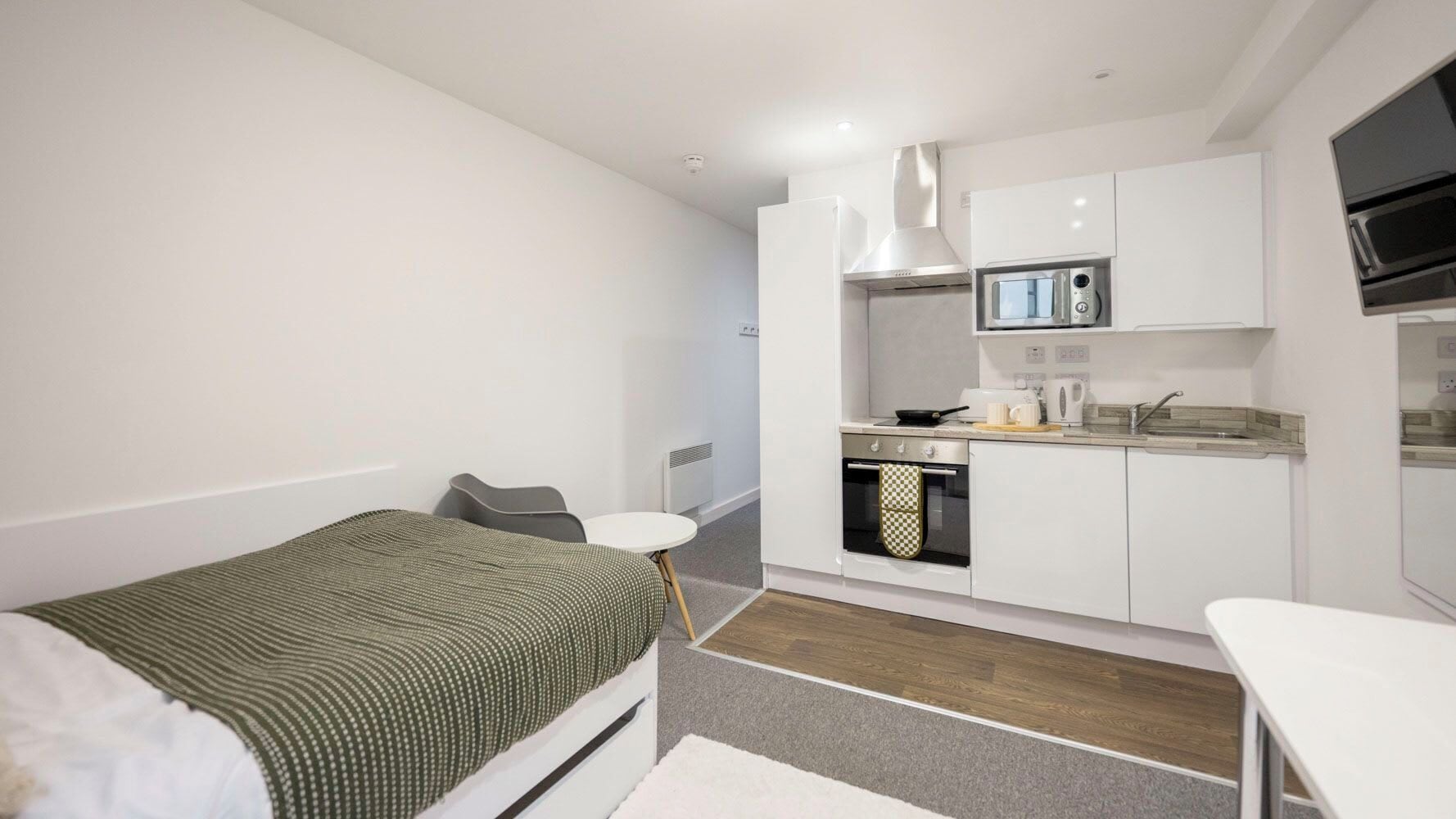 Quarto de hotel compacto com uma cama de solteiro, uma pequena cozinha, uma mesa de jantar branca e uma cadeira cinza.