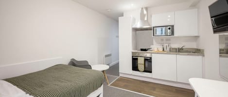 A compact hotel room with a single bed, a small kitchenette, a white dining table, and a grey chair.