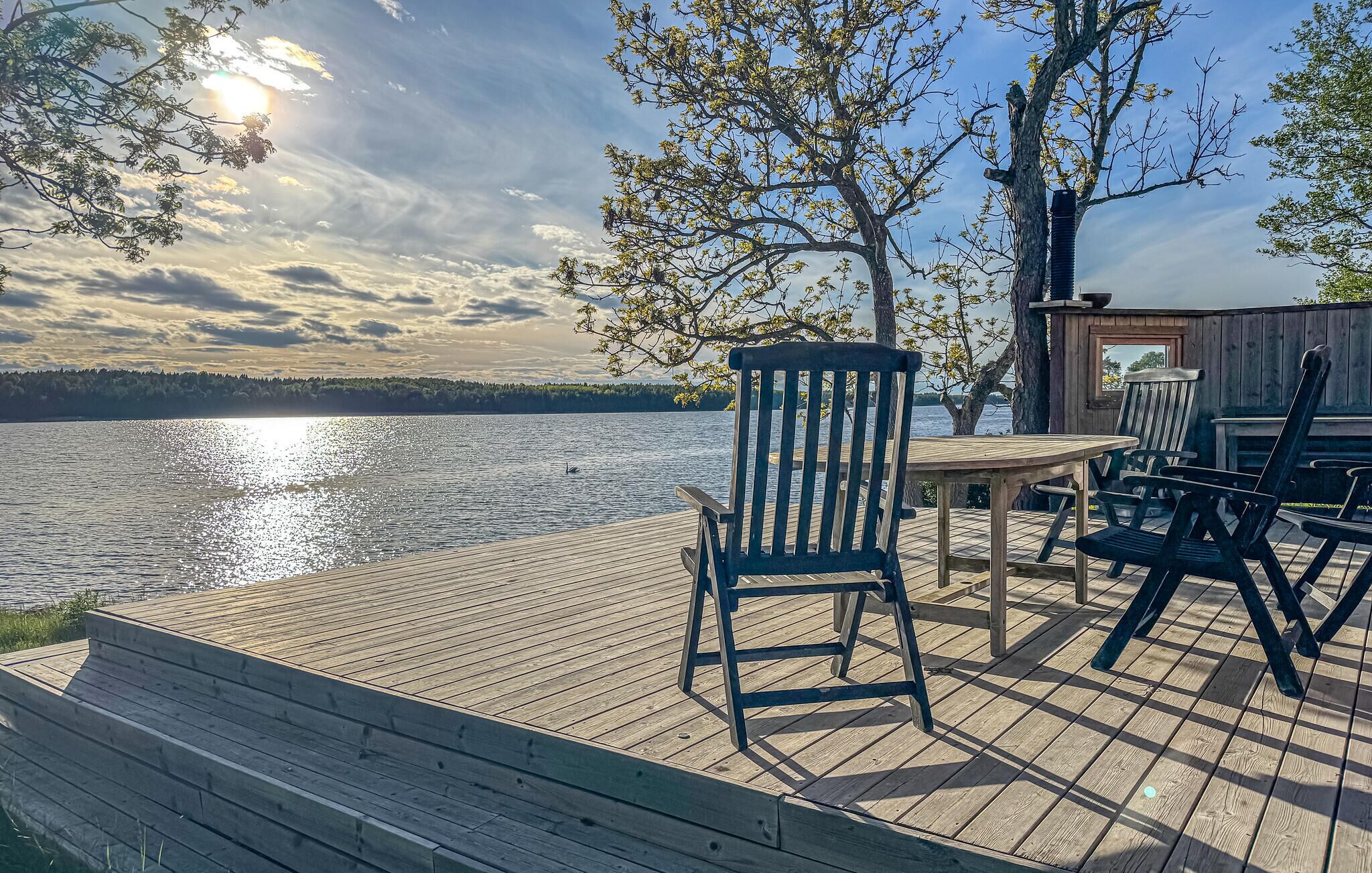 Una terraza de madera con una mesa y sillas con vistas a un lago, con árboles y una estructura de madera al fondo.