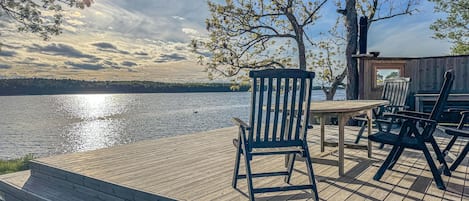 Una terraza de madera con una mesa y sillas con vistas a un lago, con árboles y una estructura de madera al fondo.
