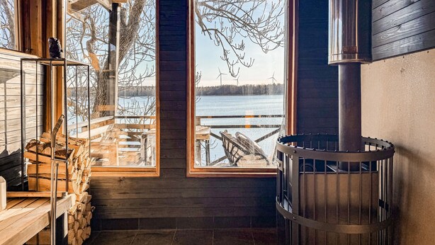 Eine Hütte mit großem Fenster mit Blick auf einen See, einem Holzofen und einem Holzstapel.