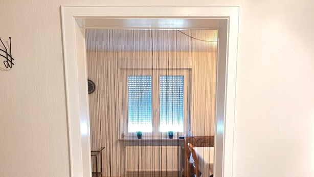 Ein Durchgang zu einem Raum mit Fliesenboden, einem Fenster mit Rollläden und einer Pflanze rechts.