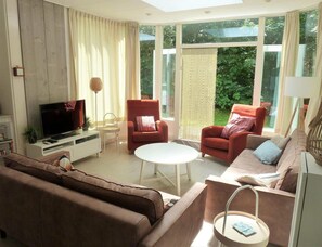 Ein Wohnzimmer mit Fernseher, zwei Sesseln, einer Couch, einem Couchtisch und einem großen Fenster mit Blick auf Grünflächen.