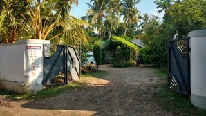 Una entrada tancada per una reixa, un camí de terra i una casa amb teulada inclinada en un entorn tropical.