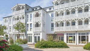 Een wit gebouw met meerdere balkons en grote ramen, omringd door groen en een grindpad.