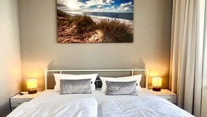 A hotel room with a bed, two bedside tables, a nightstand, a wall-mounted picture of a beach, and two pillows with text. - Stylische Ferienwohnung für Zwei mit Balkon im Seepark Sellin (Sellin)