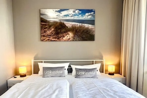 Ein Hotelzimmer mit einem Bett, zwei Nachttischen, einem Wandbild mit Strandmotiv und zwei Kissen mit Aufdruck.