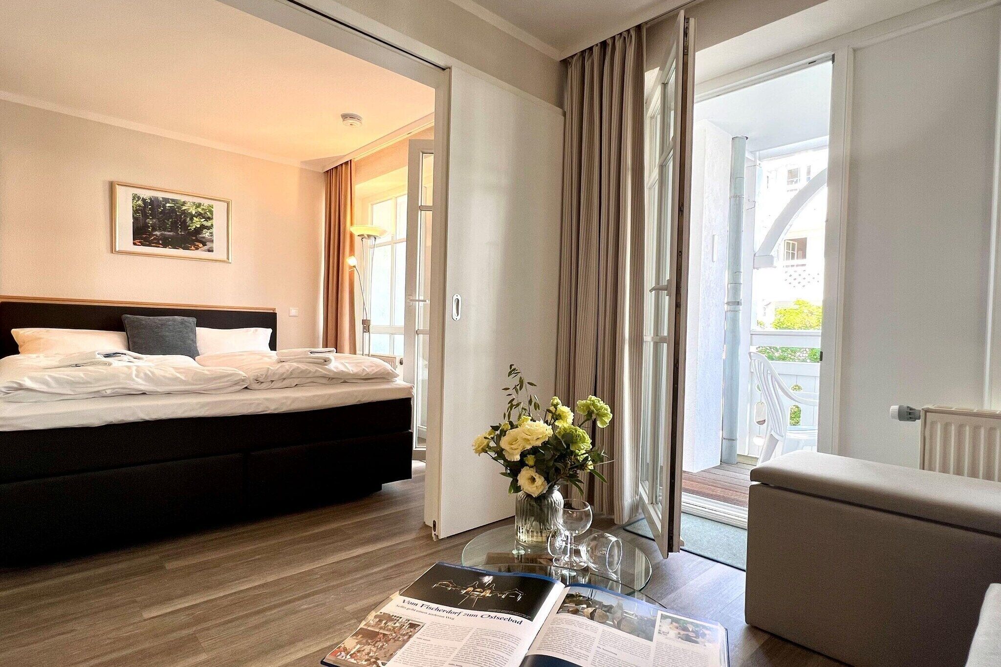 A hotel room with a bed, a glass table with a magazine, a vase with flowers, and a view of the outside.