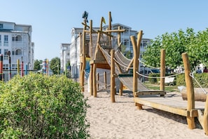 Eine hölzerne Spielgeräte-Anlage mit einer Seilbrücke und einer Kletterzone mit Netz, umgeben von Sand und Grünflächen, mit Gebäuden im Hintergrund.