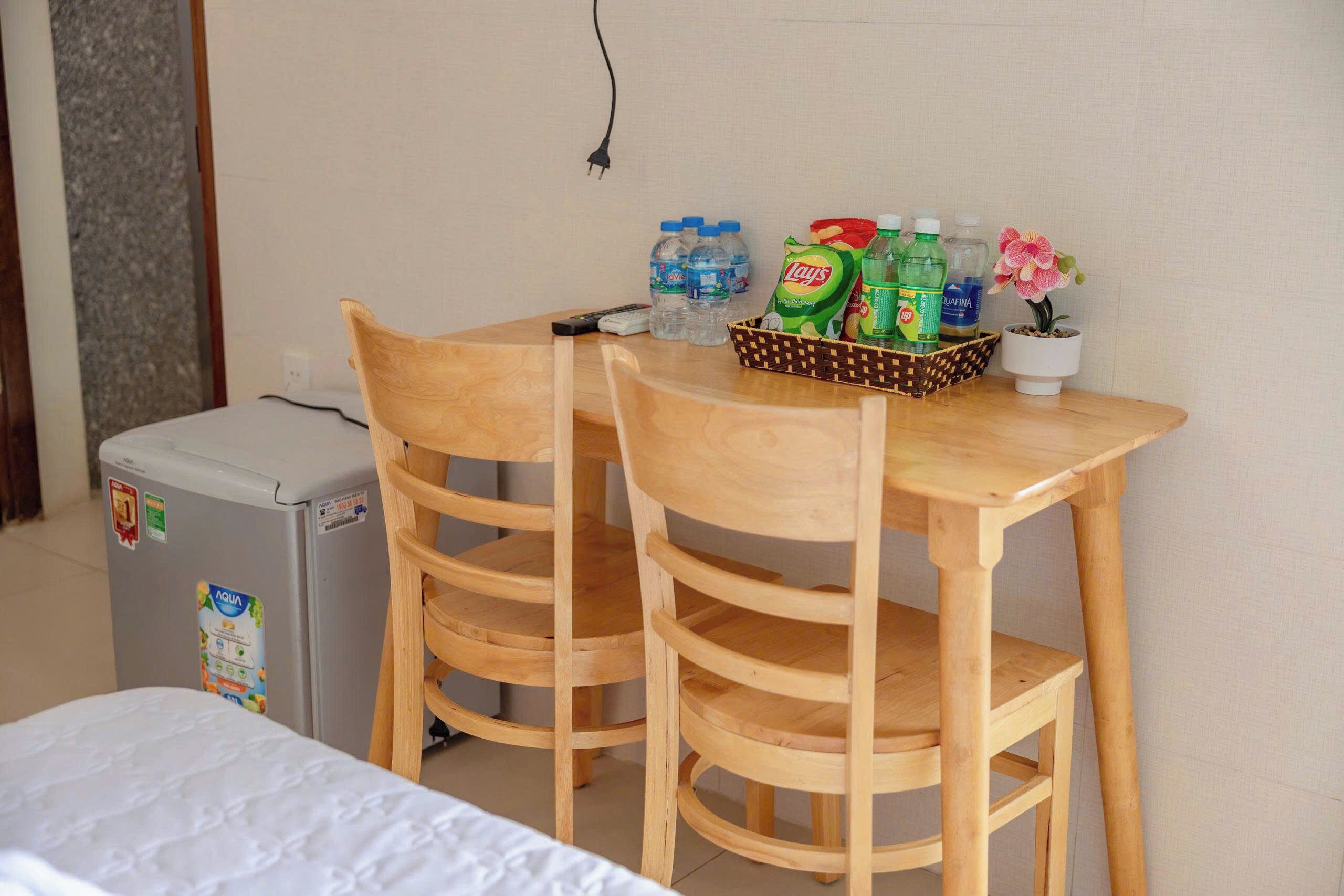 A small room with a bed, a wooden table with chairs, a refrigerator, and a basket containing toiletries.