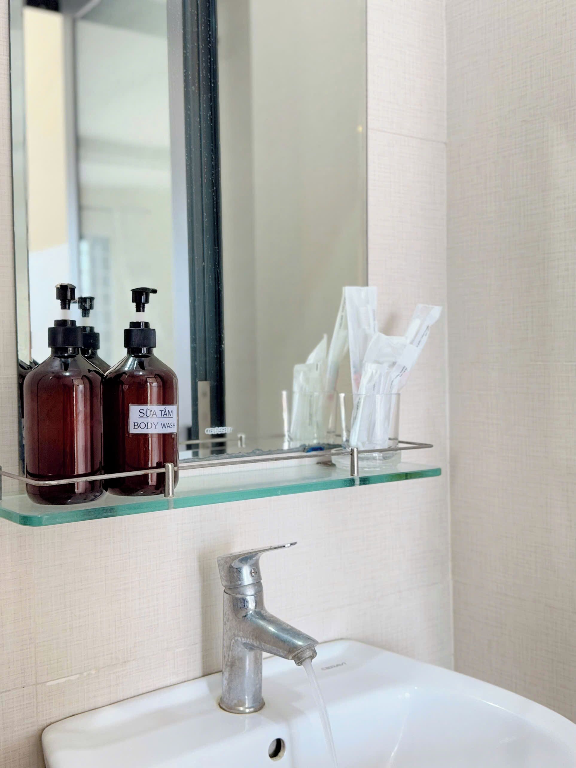 A bathroom with a white sink, a mirror, and a glass shelf holding two body wash bottles and a tissue dispenser.