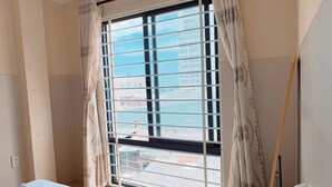Una habitación con cama, ventana con cortinas, unidad de aire acondicionado y vista a edificios.