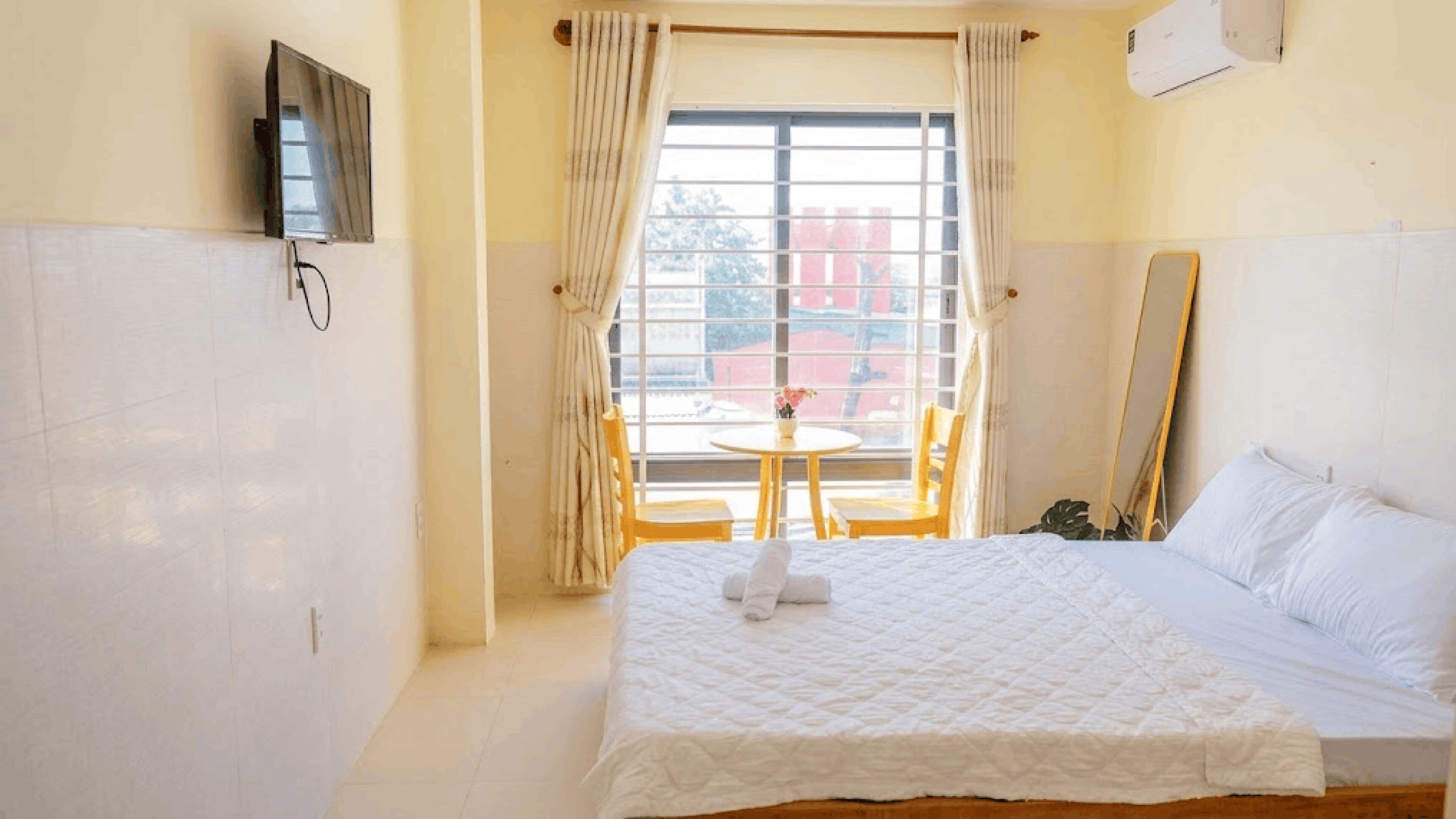 A hotel room with a bed, a TV mounted on the wall, a window with curtains, a small round table with chairs, and a mirror on the wall.