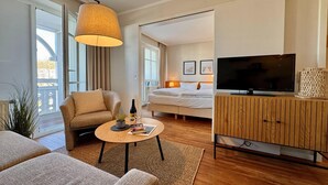 A modern hotel room with a sofa, a small round table with a bottle and glasses, a bed with a TV on a wooden cabinet, and a large window with curtains. - Stilvolle Ferienwohnung mit Balkon in Sellin (Sellin)