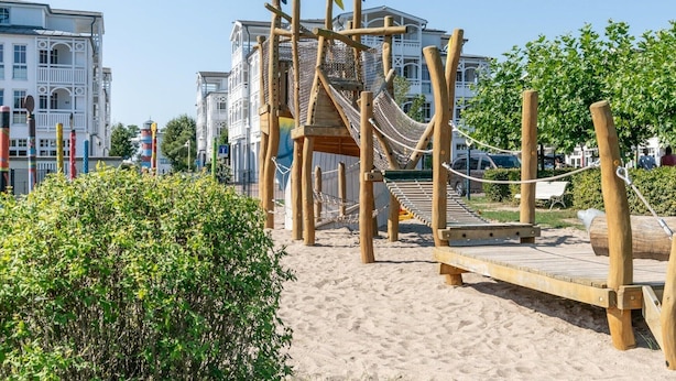 En legeplads med en tovbro og klatrestrukturer, omgivet af sand og grønt, med bygninger i baggrunden.