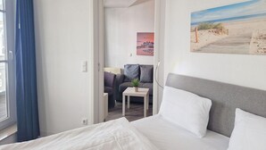 Ett hotellrum med en säng, ett litet bord med en växt, en soffa och en tavla som föreställer en strandvy.