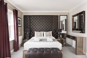 Une chambre d’hôtel moderne avec une tête de lit capitonnée, un grand lit, une table de chevet avec une lampe, un miroir et une console.