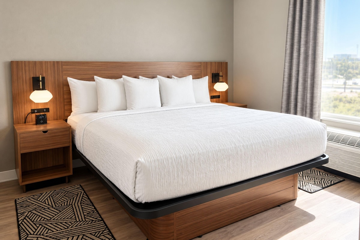 A modern hotel room with a wooden headboard, a large bed with white bedding, two bedside tables with lamps, and a view of the outdoors through a window.