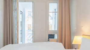 Habitación de hotel con cama, escritorio con ordenador, lámpara y ventanas amplias con cortinas.