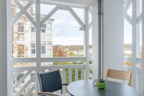 Een overdekt terras met een tafel en stoelen, uitzicht op gebouwen en bomen, en een witte balustrade.