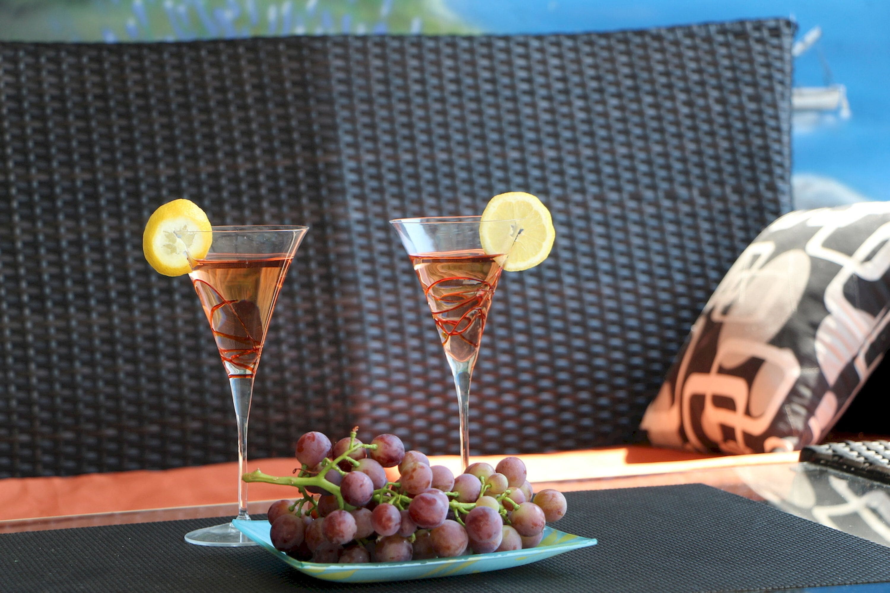 Two glasses of red beverage with lemon slices, a bunch of grapes on a plate, and a wicker sofa in the background.