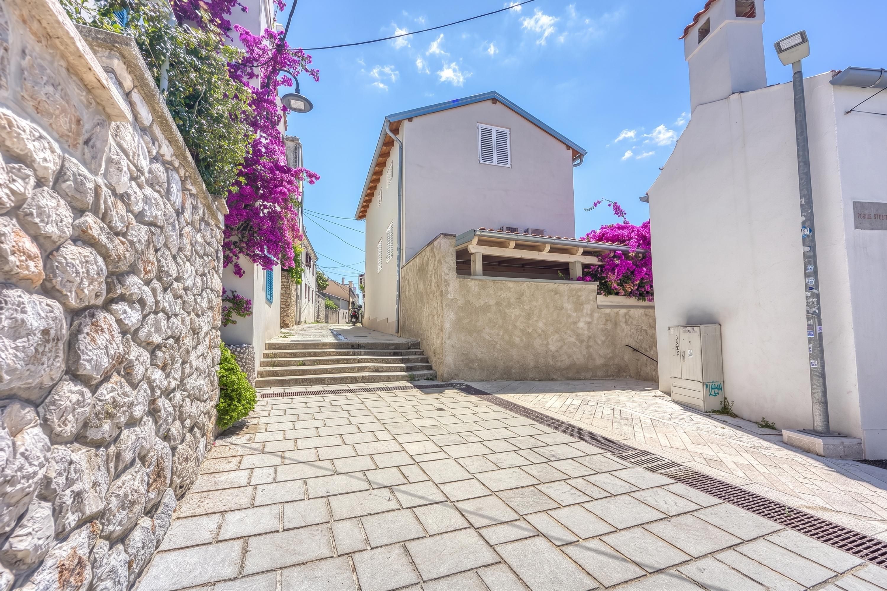 A narrow street with stone walls and a building with a wooden staircase leading to a door.
