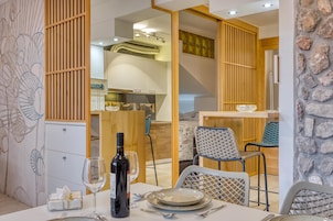 Un comedor con una mesa dispuesta para dos personas, una botella de vino y una copa. Hay sillas con un diseño tejido y una pared de piedra al fondo.
