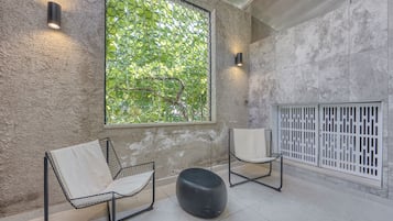 A modern interior with two metal-framed chairs, a black ottoman, a large window with a green view, and a white partition.