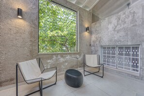 Ein modernes Interieur mit zwei stahlrahmenartigen Sesseln, einem schwarzen Hocker, einem großen Fenster mit Grünblick und einem weißen Raumteiler.