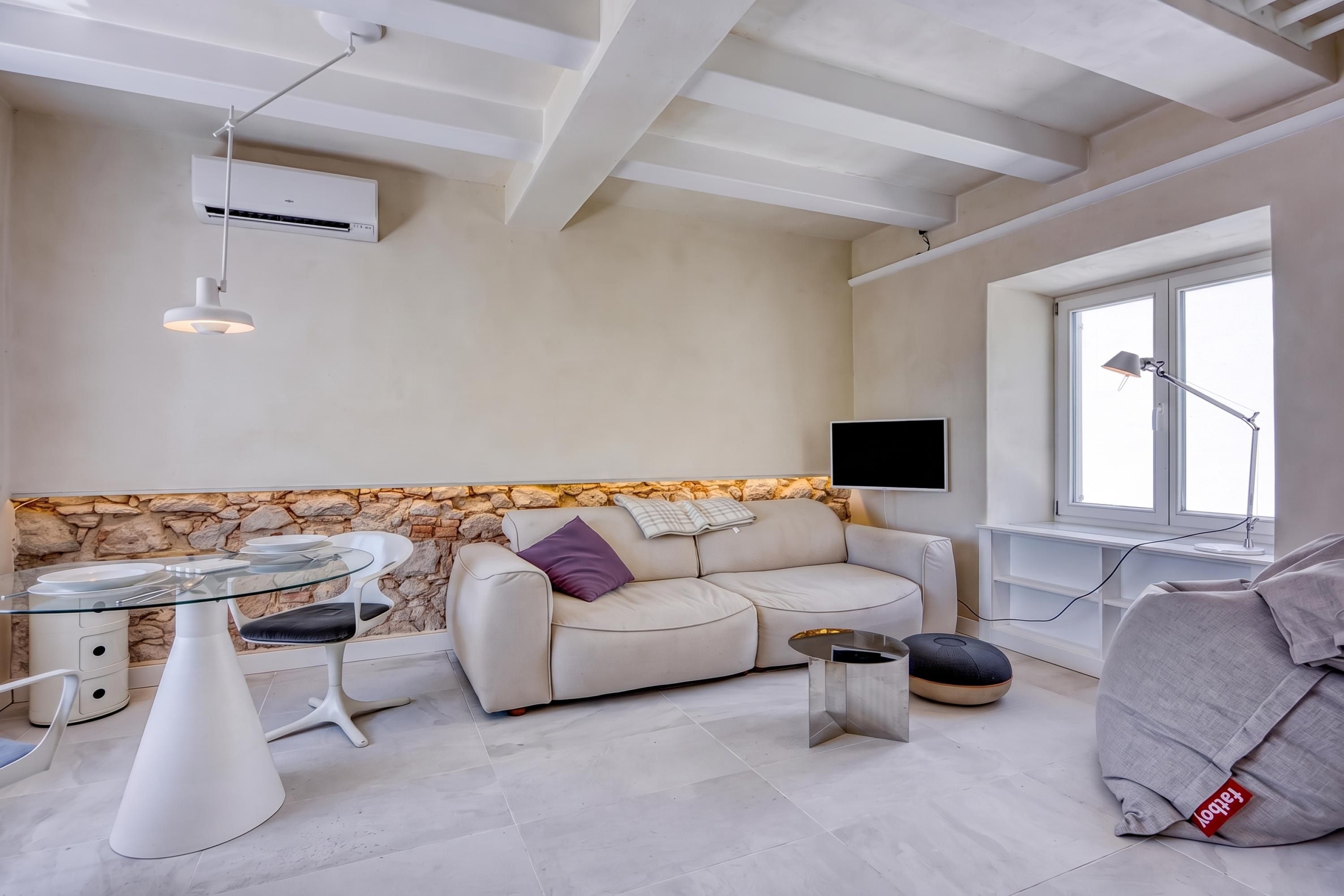 Un espacio moderno para vivir con una pared de acento de piedra, una mesa redonda de vidrio con sillas, un sofá con cojines, un televisor montado en la pared y una ventana con una lámpara de escritorio.