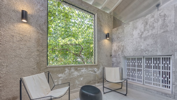 Un interior moderno con dos sillas de metal, un reposapiés negro, un ventanal con vistas a un paisaje verde y una partición blanca.