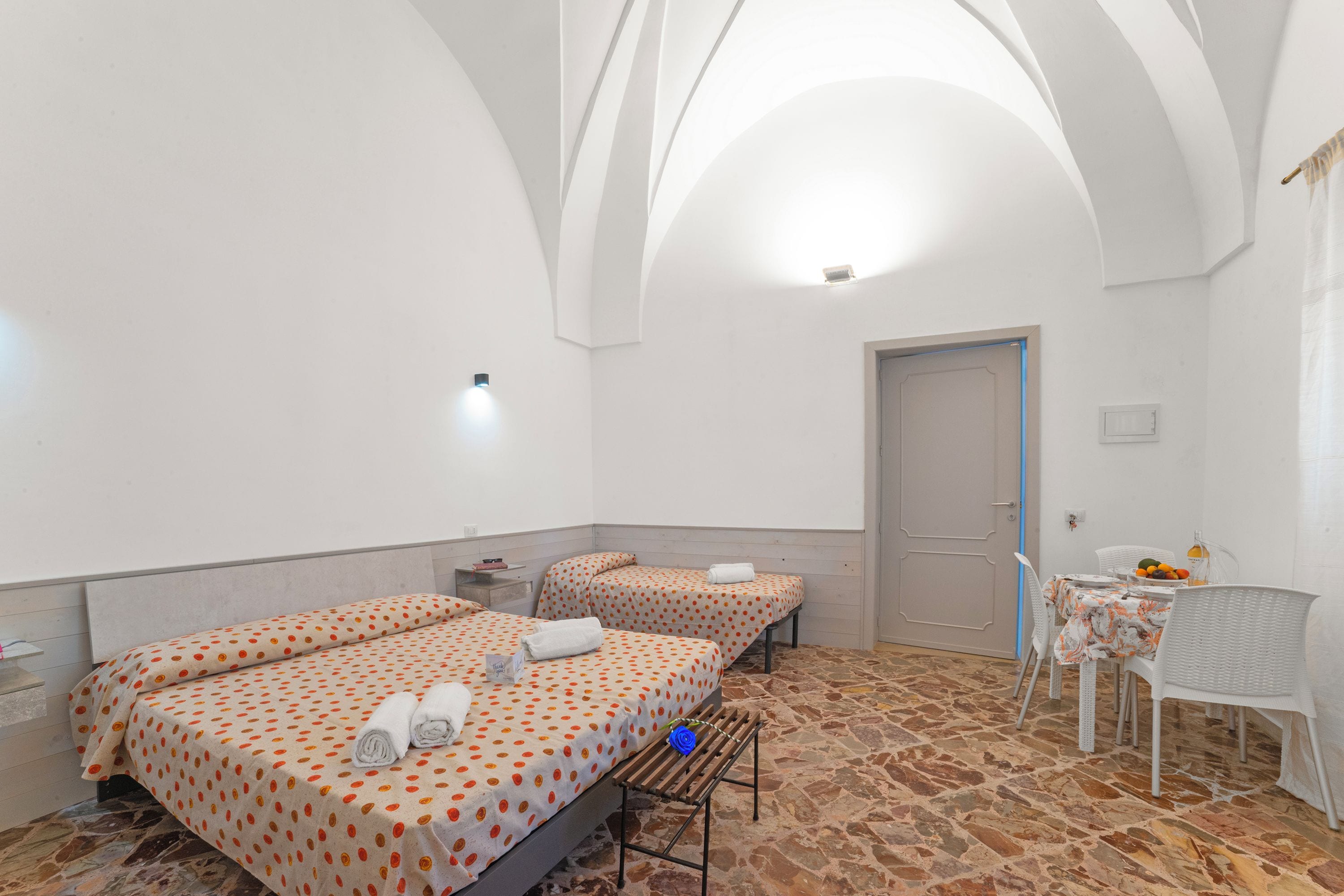 A hotel room with two beds, a small table with chairs, a bench, and a door.