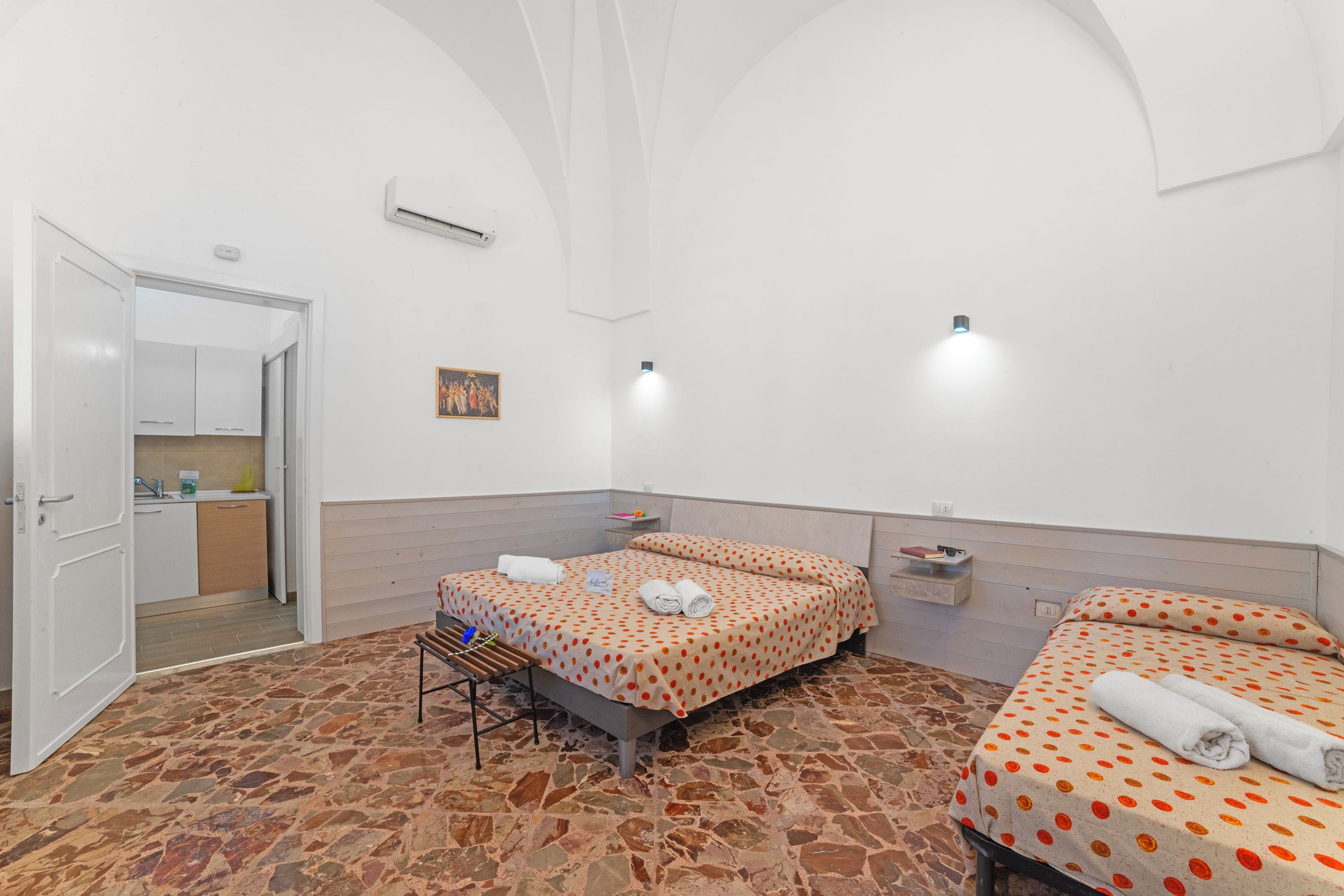 A hotel room with two beds, a small table, a bench, a painting, and a kitchen area visible through an open door.