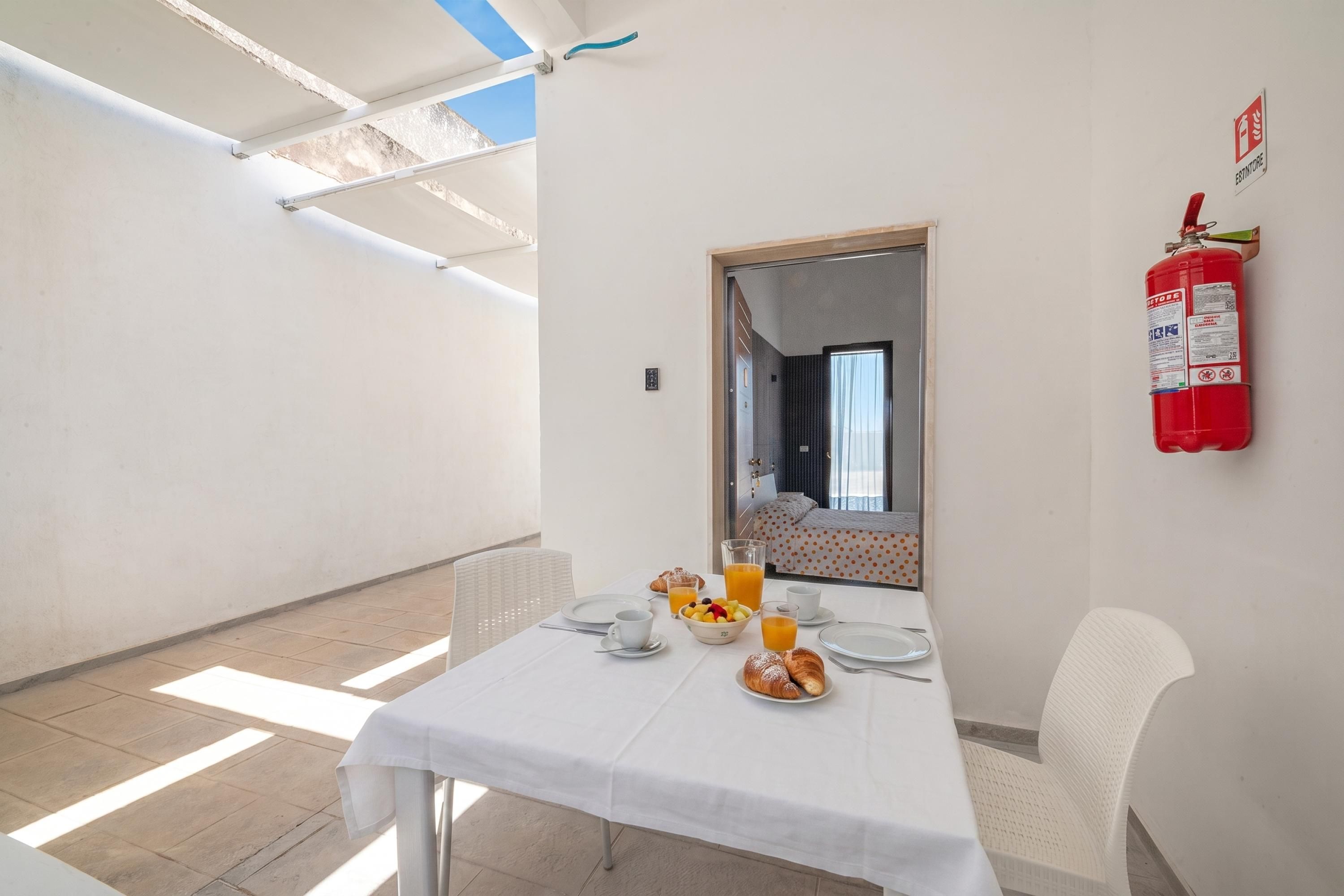 Ein Essbereich mit einem für das Frühstück gedeckten Tisch, einem an der Wand befestigten Feuerlöscher und Blick ins Zimmer durch eine Tür.