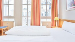 Habitación de hotel con una cama grande, una silla, una mesita de noche con lámpara y una ventana con cortinas.
